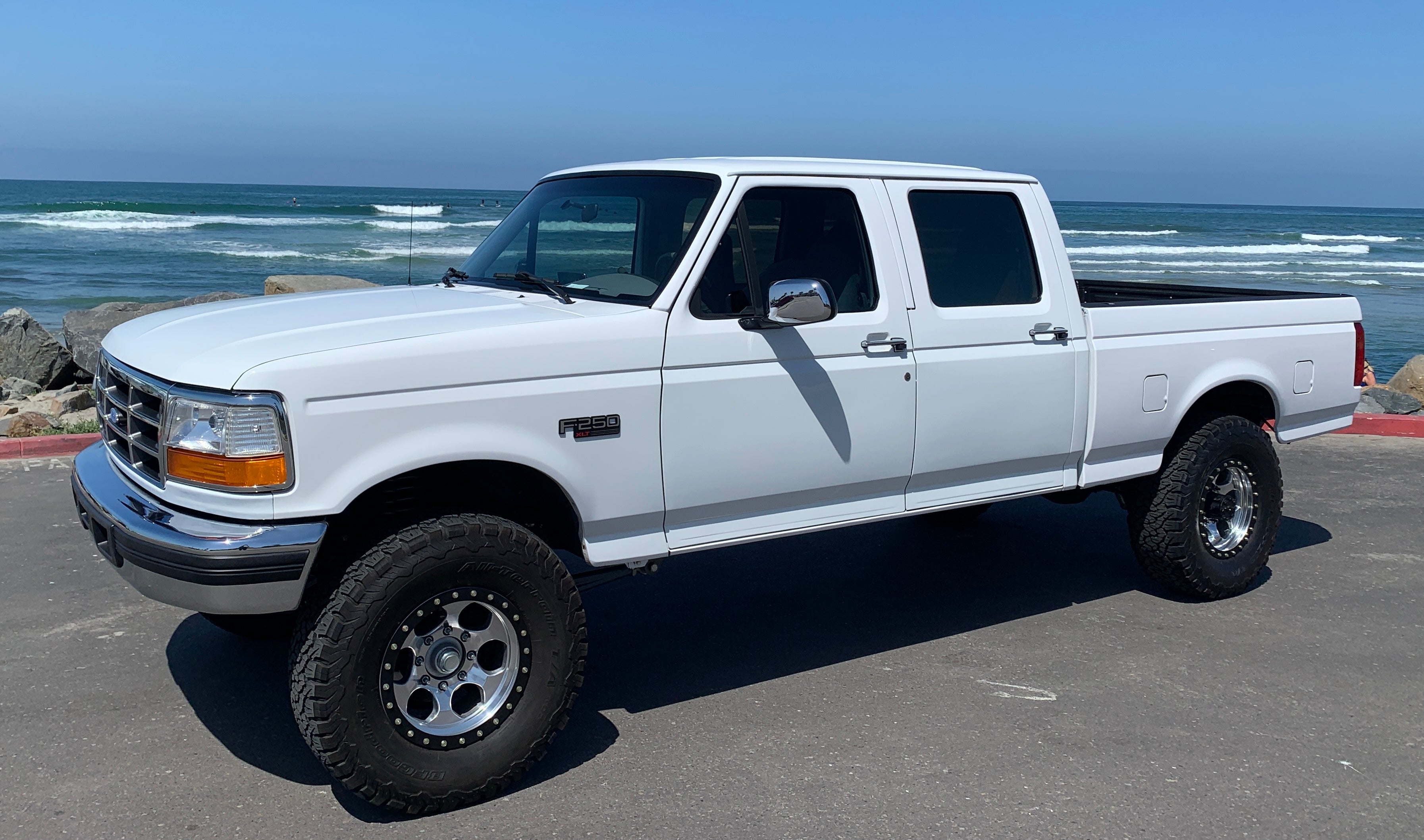 A white Ford pickup truck with a double cab and large off-road tires, parked on a paved surface with the ocean and sky in the background.